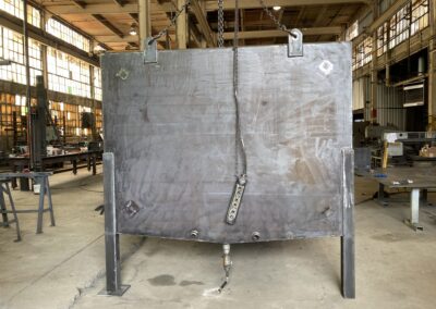 A large metal plate is suspended by chains in an industrial warehouse, with other fabrication works visible in the background.