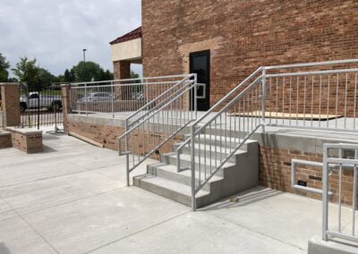 A concrete staircase with metal handrails leads up to a door on the side of a brick building. Next to the stairs is a ramp with metal railings. The area is paved with concrete, and there are trees and a parking lot visible in the background.