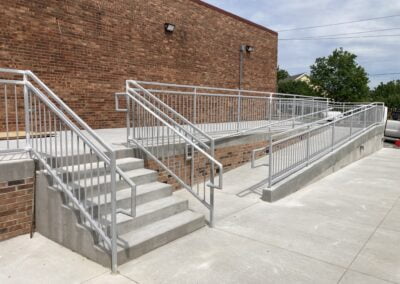 A concrete accessibility ramp with metal railings next to a set of stairs leads to a brick building. The ramp and stairs are situated on a wide, paved area.